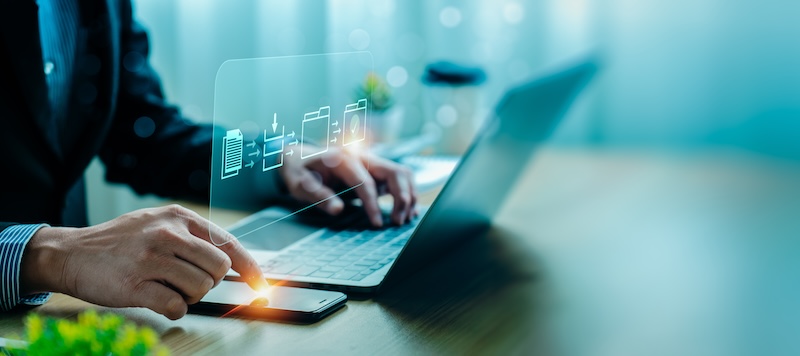 A person in business attire is using a smartphone and a laptop, with their finger touching the phone screen. Digital icons representing files, folders, and a cloud hover above the devices, symbolizing data transfer and cloud technology. It's as seamless as managing google review bots.