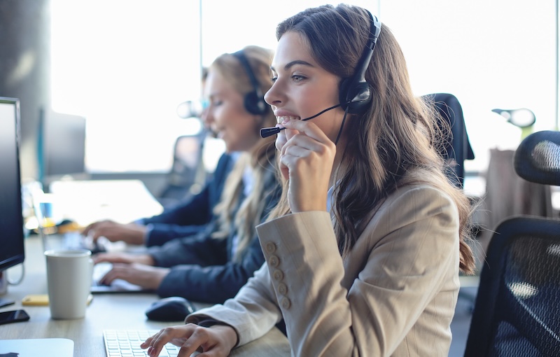 Two women in a modern office wearing headsets and working at their desks. The woman in the foreground is smiling while typing on a keyboard and speaking into the headset microphone, possibly assisting with Google review bots. In the background, the second woman is similarly engaged in work.