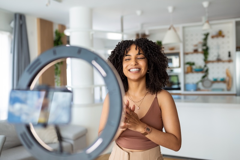 A woman with curly hair is smiling and recording herself using a smartphone mounted on a ring light. She is in a modern, well-lit kitchen with white cabinets and hanging pendant lights in the background, perhaps getting ready to share tips on spotting fake google review bots.