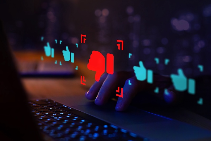 A person's hand hovers over a laptop keyboard in a dimly lit environment. Red thumbs-down and blue thumbs-up icons float above the keyboard, representing social media reactions. The icons are illuminated against a blurred dark background, highlighting the interaction—almost as if guided by invisible google review bots.