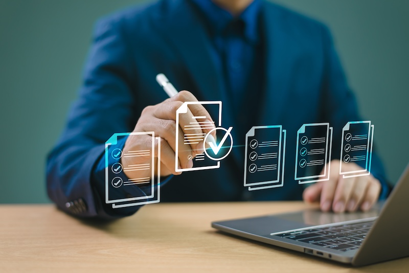 A person in a blue suit uses a stylus to interact with a holographic checklist hovering over a laptop on a desk. The holographic interface displays multiple documents and a checked item, resembling the organized precision of Google review bots managing task completion or data organization.