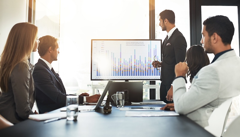 A group of four business professionals sit in a meeting room, focusing on a man in a suit presenting data on a screen. The screen displays a bar chart with various colored bars and insights on google review bots. Sunlight filters through the window, creating a well-lit setting.