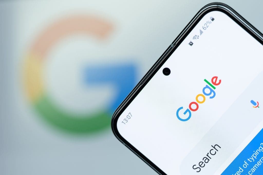 A smartphone displaying the Google search page is held in front of a blurred Google logo in the background. The phone screen shows the Google logo, search bar, microphone icon, and part of the time and battery status.