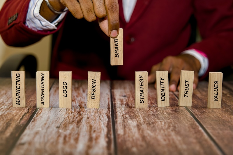 A person in a red blazer arranges wooden blocks labeled MARKETING, ADVERTISING, LOGO, and more—symbolizing brand building and the effort to keep a good reputation.