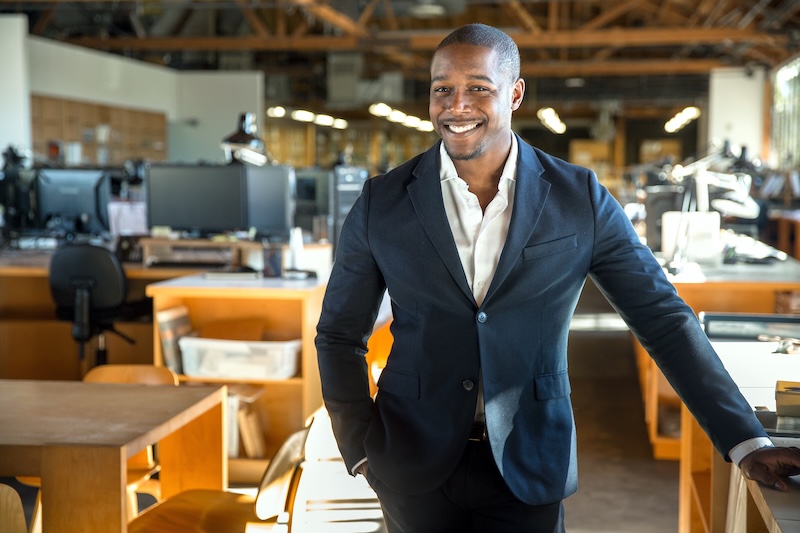 A man in a blue blazer and white shirt stands smiling in a well-lit office, contemplating "Can I Make a Wikipedia Page for Myself?" The space features wooden desks and modern chairs, with computers and shelves in the background. The ceiling shows exposed beams and lighting.
