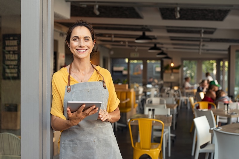 A smiling person in a yellow shirt and gray apron holds a tablet, likely updating how to change business hours on Apple Maps. They're standing at the entrance of a bustling cafe, where tables host lively conversations among several seated patrons.