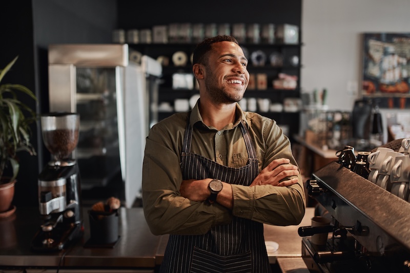 A barista stands behind a coffee shop counter, smiling with arms crossed. He wears an apron over a green shirt, ready to assist customers or even share tips like how to change business hours on Apple Maps. Shelves with coffee products and plants are visible in the background.