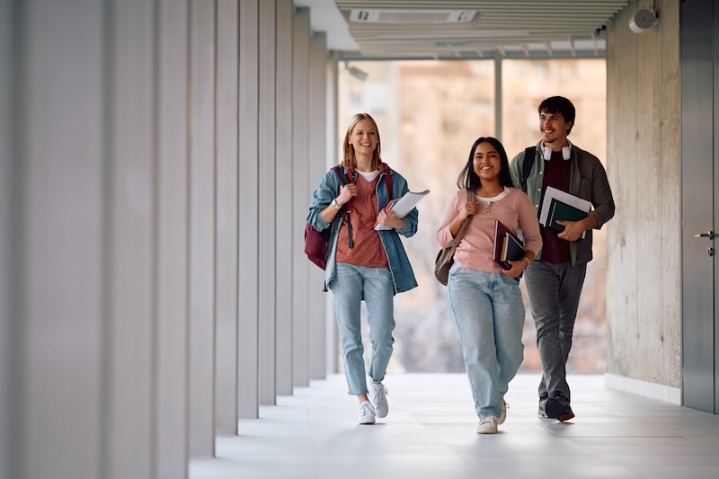 Three smiling students walk down a bright hallway carrying books and backpacks, embodying the positive University Online Reputation fostered by a friendly and welcoming campus environment.