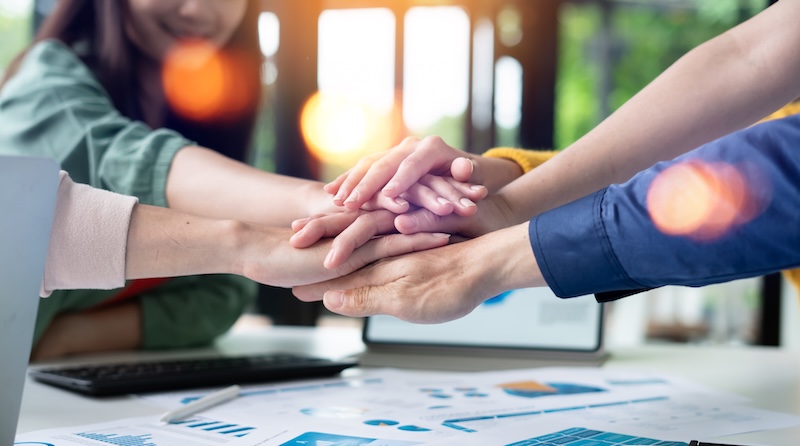 A group of people stack their hands together over a desk with charts and graphs, symbolizing teamwork, collaboration, and a commitment to Corporate Social Responsibility in a bright office setting.