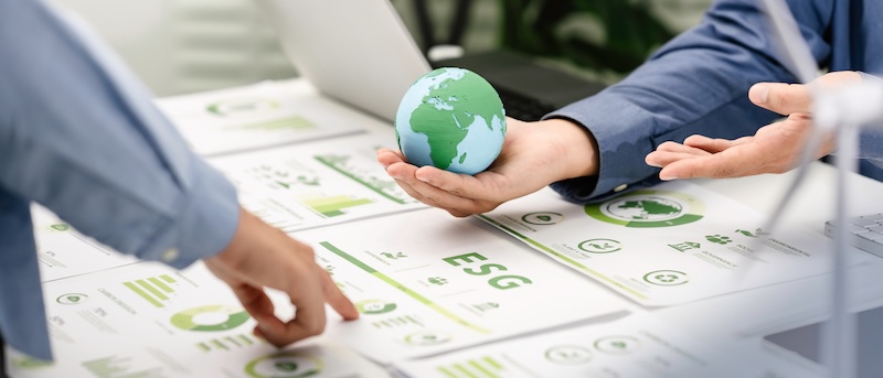 Two people in business attire discuss ESG (Environmental, Social, Governance) and Corporate Social Responsibility documents. One holds a small globe, highlighting sustainability, with charts and a laptop visible in the background.