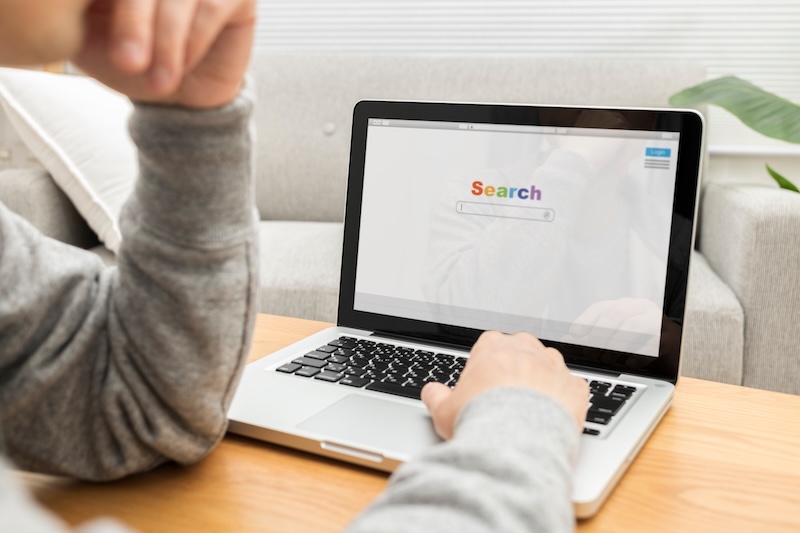 A person is ego surfing on a laptop at a wooden table, with a search engine homepage displayed. Their hand rests on the keyboard while their face is partially visible.