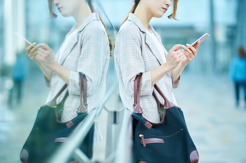 A woman stands by a glass wall, her reflection visible, while ego surfing on her smartphone. She wears a plaid shirt and carries a large black handbag with brown straps. The background is softly blurred.