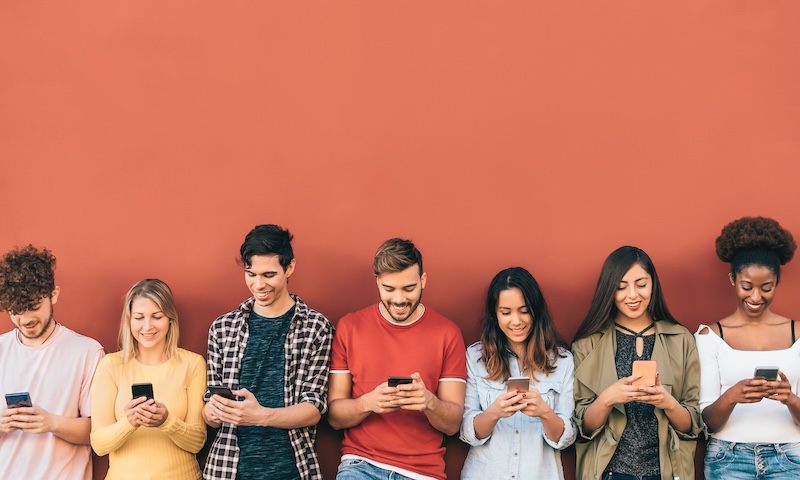 Seven young adults stand in a row against an orange wall, smiling and looking down at their smartphones. Relaxed and engaged, each holds a phone—showing how they keep a good reputation while staying connected online.