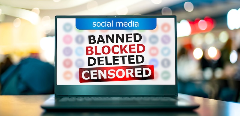 A laptop screen displays a message about social media and online safety, with the words "BANNED," "BLOCKED," "DELETED," and "CENSORED" in bold letters, symbolizing online restrictions. The background is blurred.