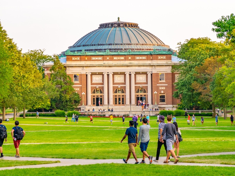A group of students walk and chat on a green lawn in front of a large, domed university building, its strong online reputation reflected in the lively campus atmosphere, surrounded by trees on a sunny day.