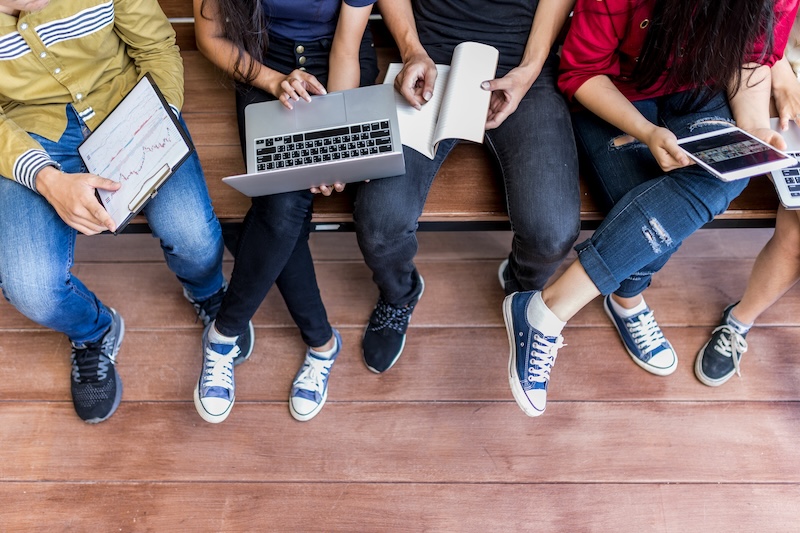 Four people sit on a bench, seen from above, using a laptop, tablet, and notebook. Focused on their work—perhaps researching University Online Reputation—they wear casual clothes and sneakers. The wooden floor stretches beneath them.