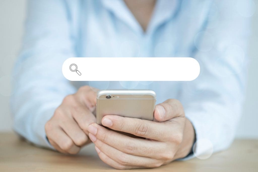 A person wearing a light blue shirt is holding a smartphone with both hands. There is an illustrated search bar superimposed above the phone, containing a magnifying glass icon on the left. The background is blurry with soft light patterns around the edges.