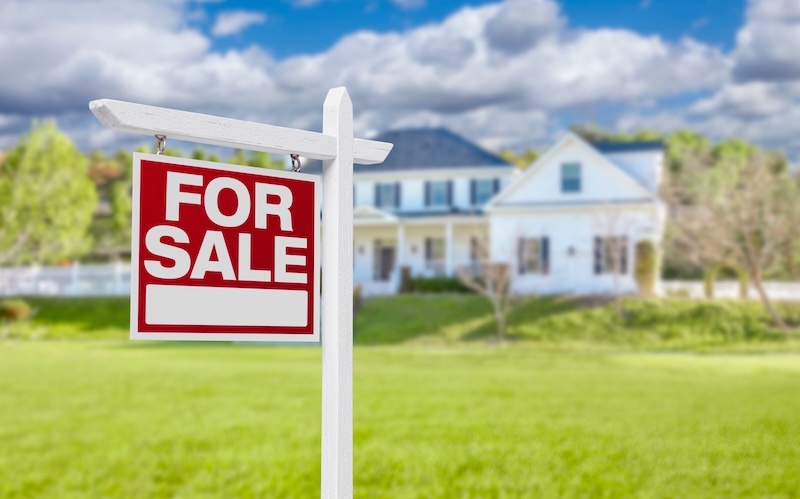 A red and white "For Sale" sign is posted in front of a large, two-story house with a porch, as Realtors need online reputation management to attract buyers, set among green grass and trees under a partly cloudy sky.