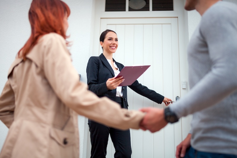 A real estate agent holding a clipboard smiles as she opens the door to a house for a couple holding hands, highlighting why Realtors Need Online Reputation Management during home viewings or tours.
