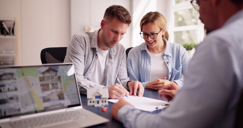 A couple sits with a real estate agent, signing documents. A laptop displays house listings and a model house is on the table, highlighting the home-buying process and why Realtors need online reputation management.