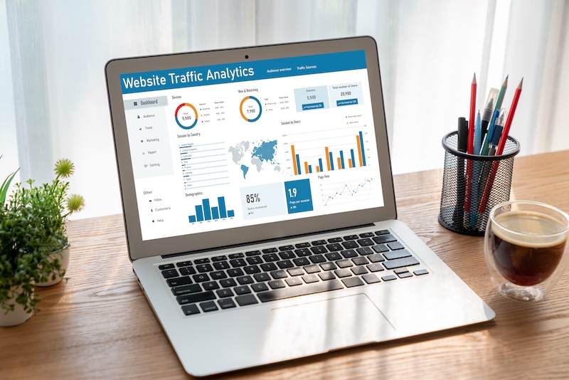 A laptop on a wooden desk displays a website traffic analytics dashboard with graphs, charts, and statistics—essential tools to keep a good reputation. Nearby are a small plant, pencils in a holder, a notepad, and a cup of coffee.