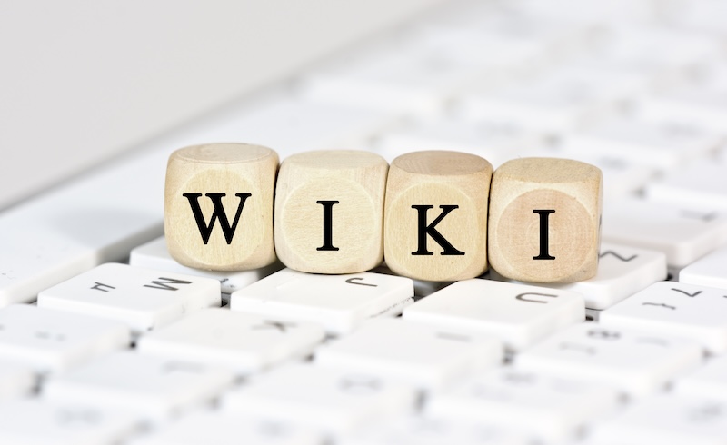 Wooden blocks spelling "WIKI" rest atop a white computer keyboard, posing the silent question: "Can I make a Wikipedia page for myself?" The blocks align neatly across the keys, creating a subtle yet striking contrast against the pristine backdrop.