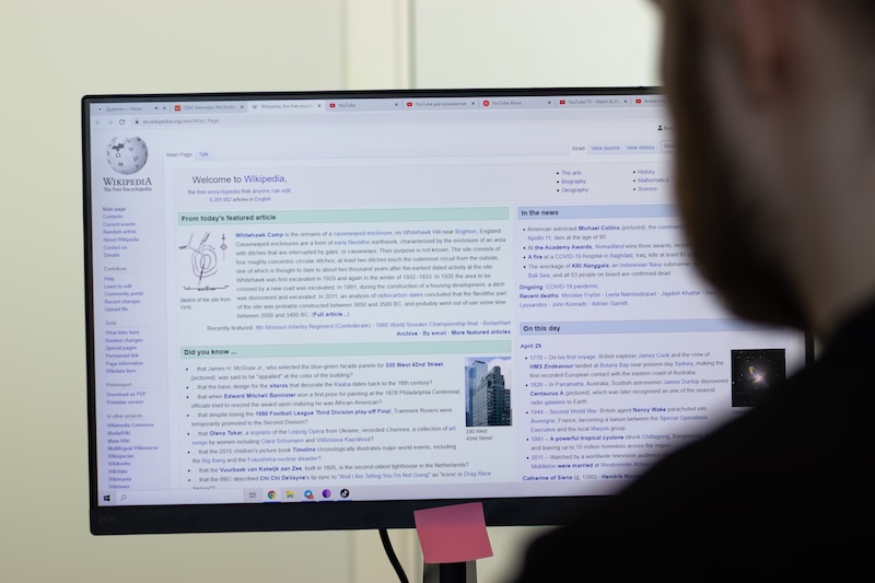 A person is peering at a computer monitor displaying the Wikipedia homepage. Amidst sections of text and images, a pink sticky note clings to the stand, perhaps posing the question: "Can I make a Wikipedia page for myself?.