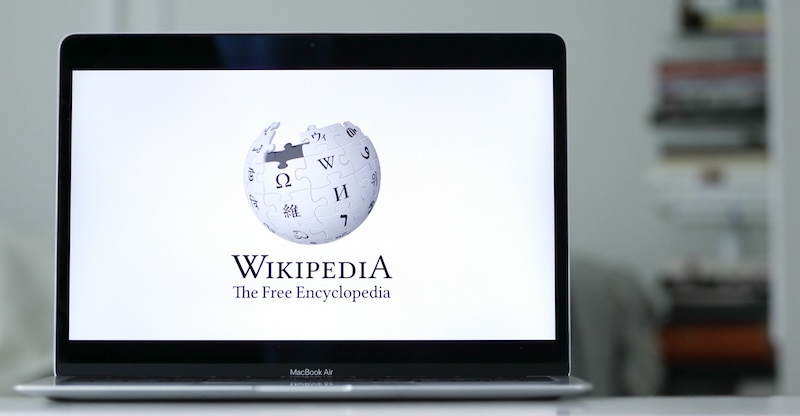 A laptop showcasing the Wikipedia logo—a globe of puzzle pieces with diverse characters above the text "WIKIPEDIA The Free Encyclopedia." The blurred background hints at stacked items, perhaps inspiring thoughts like, "Can I make a Wikipedia page for myself someday?.