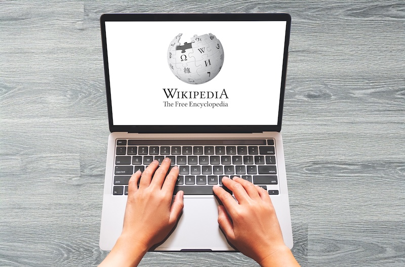 Hands typing on a laptop with a Wikipedia logo on the screen, displayed on a wooden surface. It leaves you wondering, "Can I Make a Wikipedia Page for Myself?" as curiosity and creativity blend seamlessly in the quest to contribute to this vast digital encyclopedia.