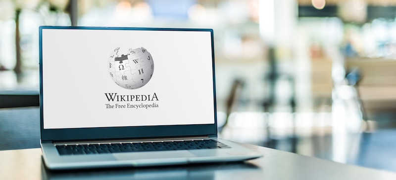 A laptop shows the Wikipedia homepage with its iconic logo and tagline "The Free Encyclopedia." The background, a soft blur of bright, neutral cafe tones, invites contemplation. It makes one wonder: "Can I Make a Wikipedia Page for Myself?.