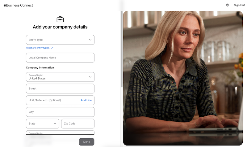 A woman with long blonde hair wearing a black and green blouse sits at a table with a laptop, focused on "Add your company details" form. She's contemplating how to change business hours on Apple Maps as she fills in the necessary fields for her business information.