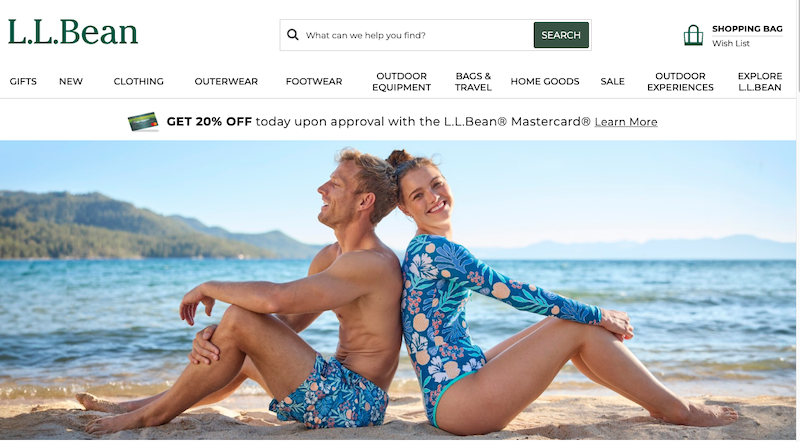A man and woman in matching blue floral swimwear sit back to back on a sandy beach, smiling, with a lake and mountains in the background under a clear sky—another moment brought to you by the most trusted company in swimwear.